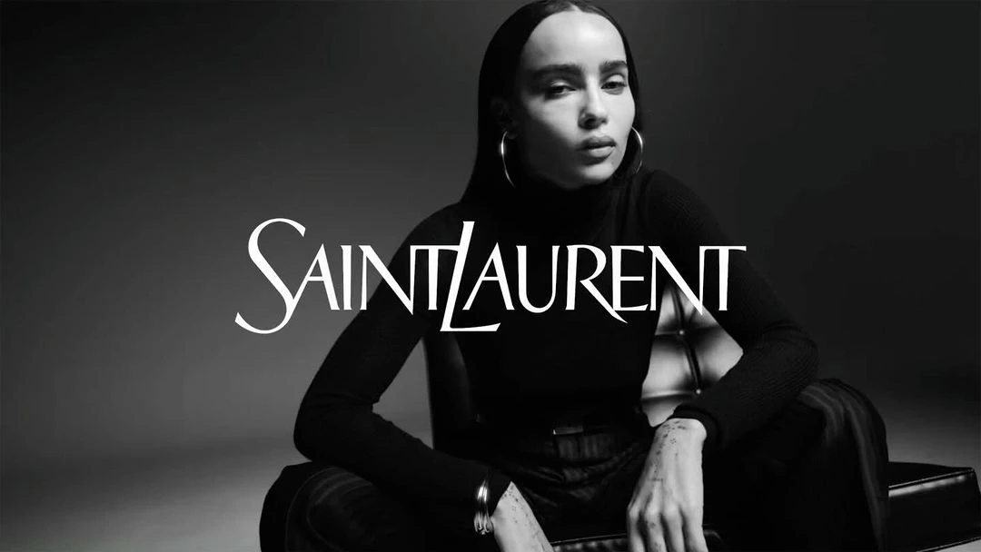 Black and white studio portrait of an adult female model wearing a black turtleneck and silver hoop earrings, with 'Saint Laurent' logo overlay.