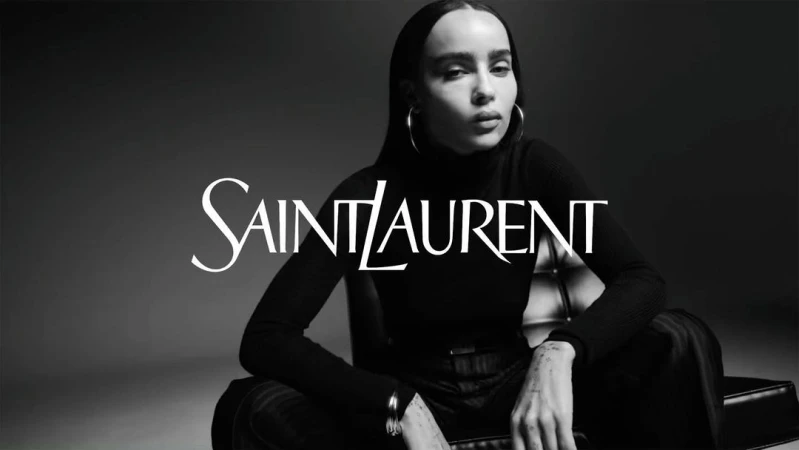 Black and white studio portrait of an adult female model wearing a black turtleneck and silver hoop earrings, with 'Saint Laurent' logo overlay.