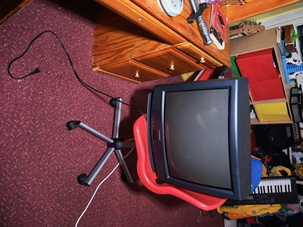 Vintage television sitting on a red chair in a room with a wooden desk and patterned carpet. The room appears to be a home or office.