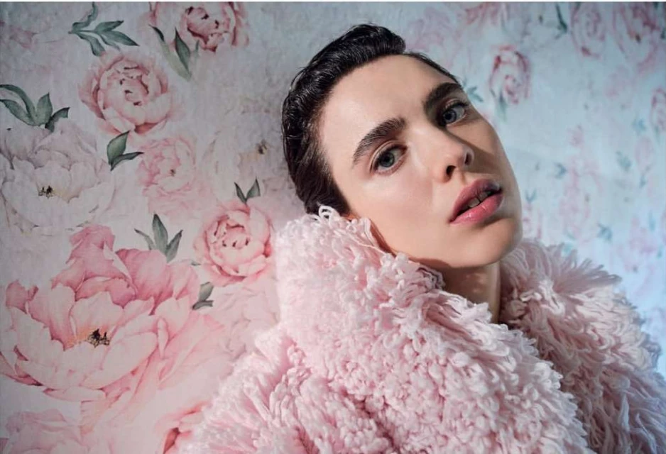 Portrait of a fair-skinned adult female with blue eyes wearing a pink furry jacket against a floral wallpaper background.