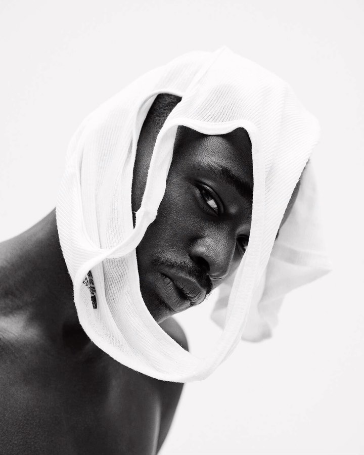 Monochrome portrait of a Black man wearing a white towel on his head, looking at the camera against a bright white background.