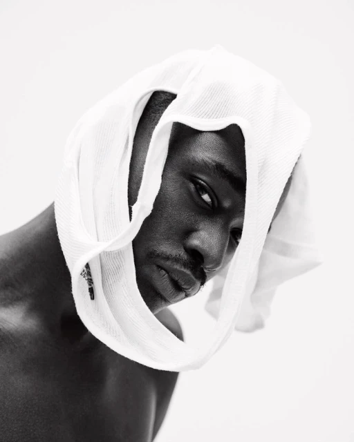 Monochrome portrait of a Black man wearing a white towel on his head, looking at the camera against a bright white background.