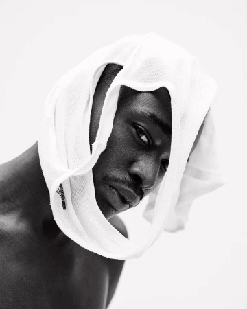 Monochrome portrait of a Black man wearing a white towel on his head, looking at the camera against a bright white background.