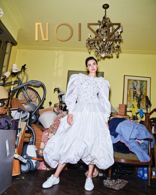 Woman in white gown poses indoors amid cluttered room with vintage furniture and chandelier, bathed in bright, diffused light.