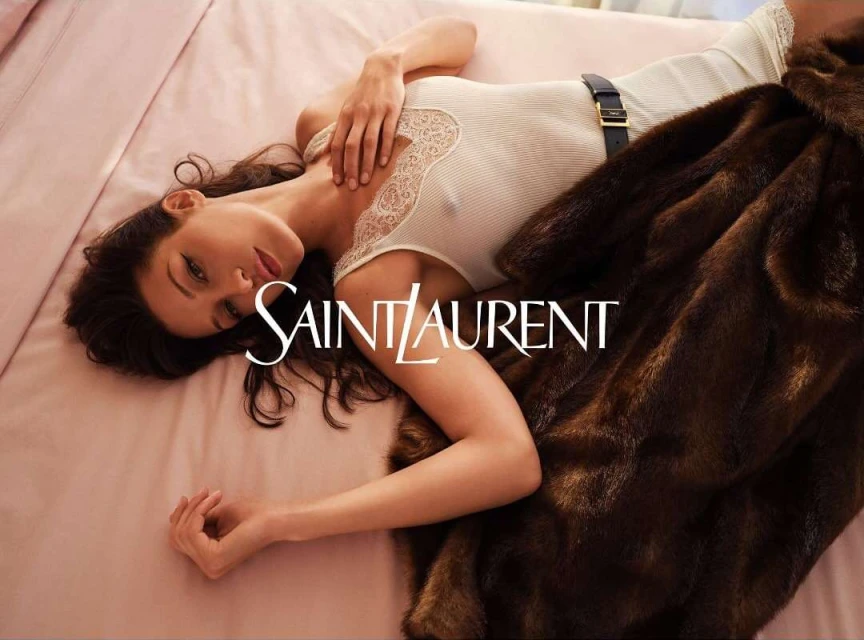 Adult woman poses in a white, lace-trimmed tank top and black belt while lying on a pink bed next to a dark brown fur coat.