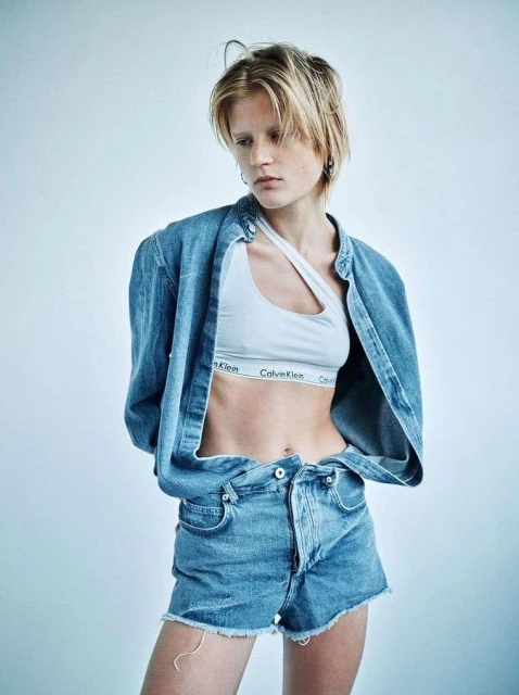 Blonde woman in a Calvin Klein bra top and denim shorts and jacket posing in a photo studio with a neutral background and soft lighting.