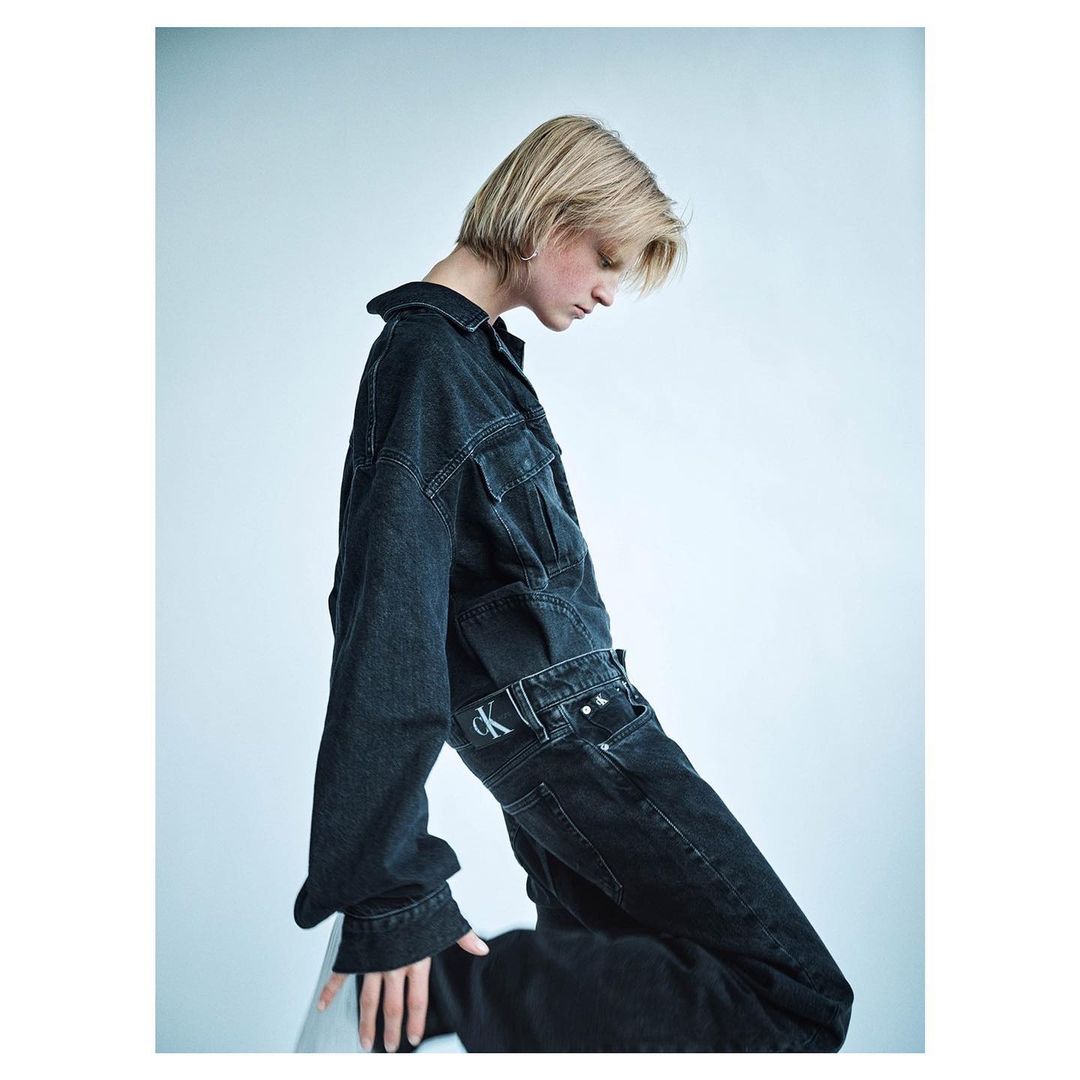 Model with blonde hair wearing dark denim jacket and matching jeans against pale studio background