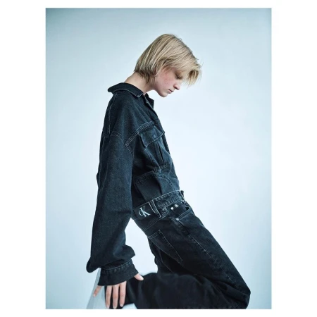 Model with blonde hair wearing dark denim jacket and matching jeans against pale studio background