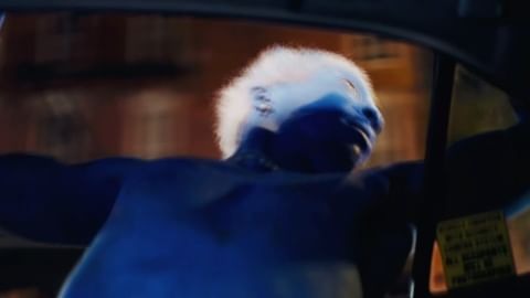 A man with bright white hair looks out the window of a car at night with visible city lights in the background. The scene is color graded with blue.