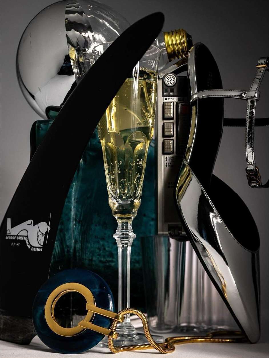 Still life shot of a champagne flute, metallic high heel shoe, bottle opener, and a lightbulb, all grouped together indoors.