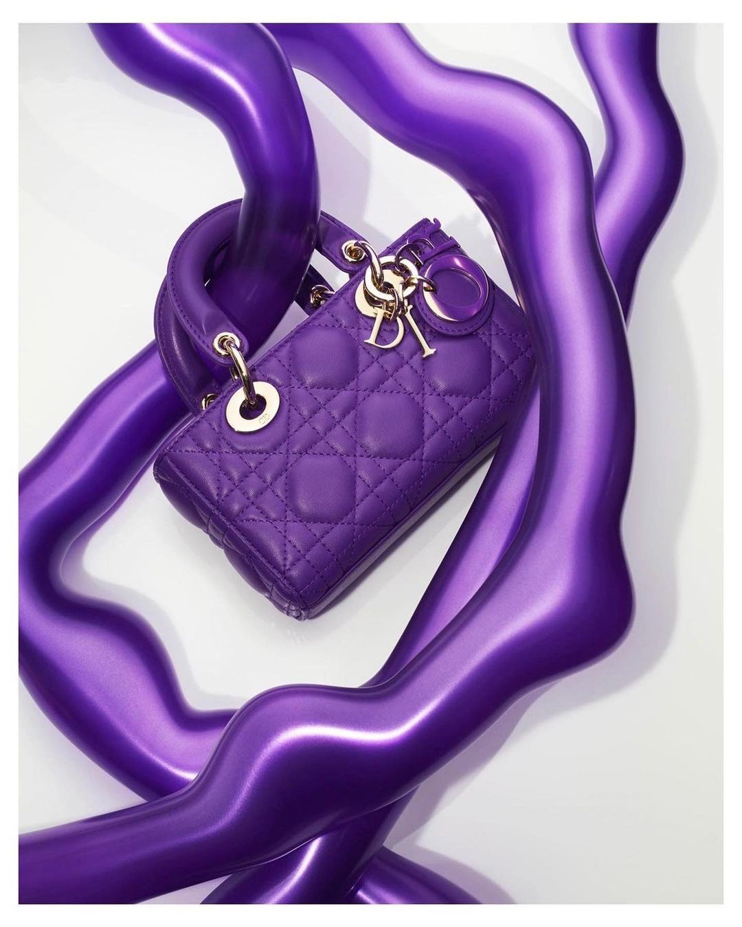 Elegant, quilted Lady Dior handbag in vibrant purple with gold hardware, gracefully intertwined with glossy, abstract purple forms.