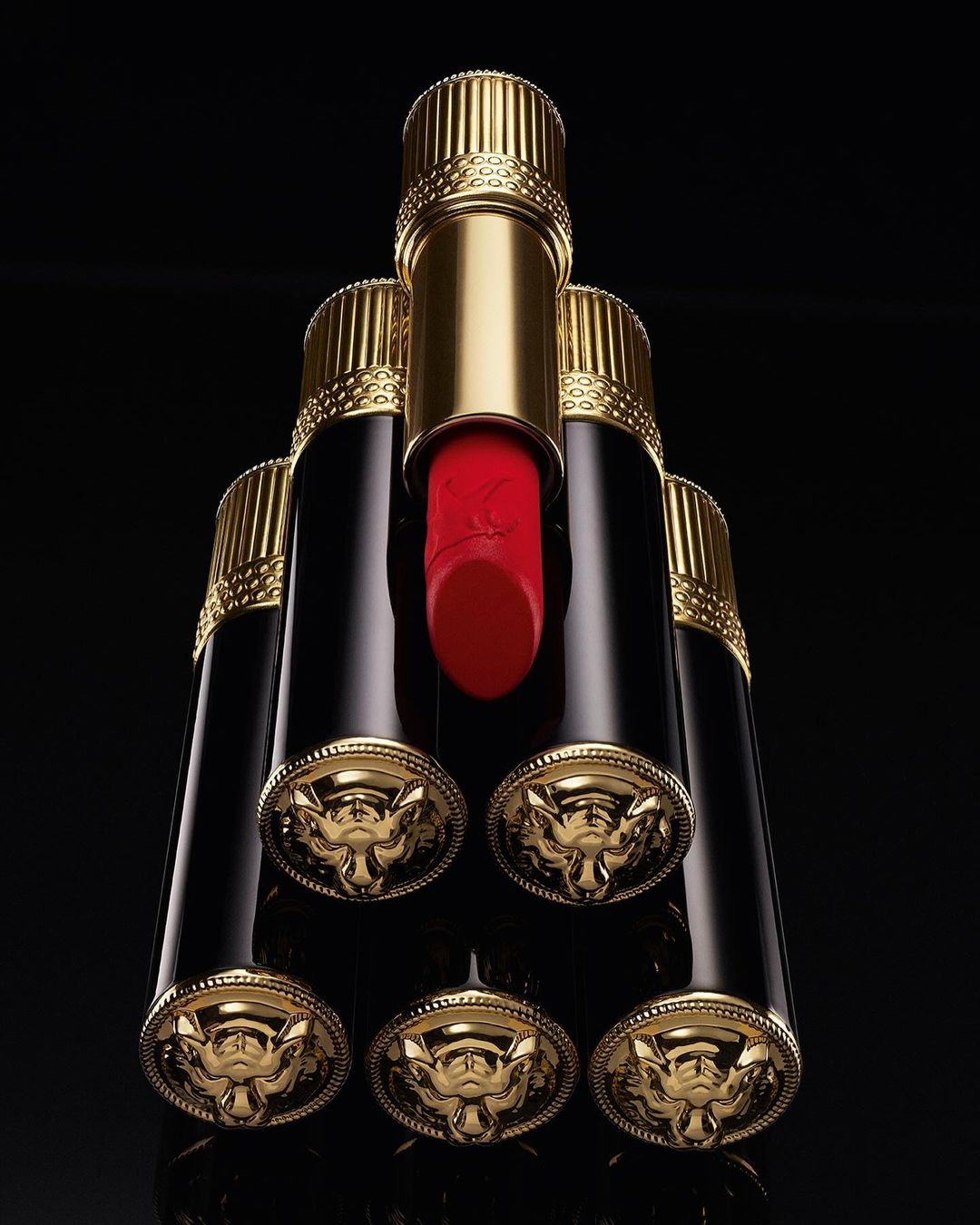 Luxury beauty still life: A pyramid arrangement of gold and black lipsticks with red visible on the top one against a black background.