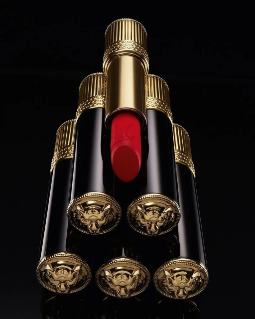 Luxury beauty still life: A pyramid arrangement of gold and black lipsticks with red visible on the top one against a black background.