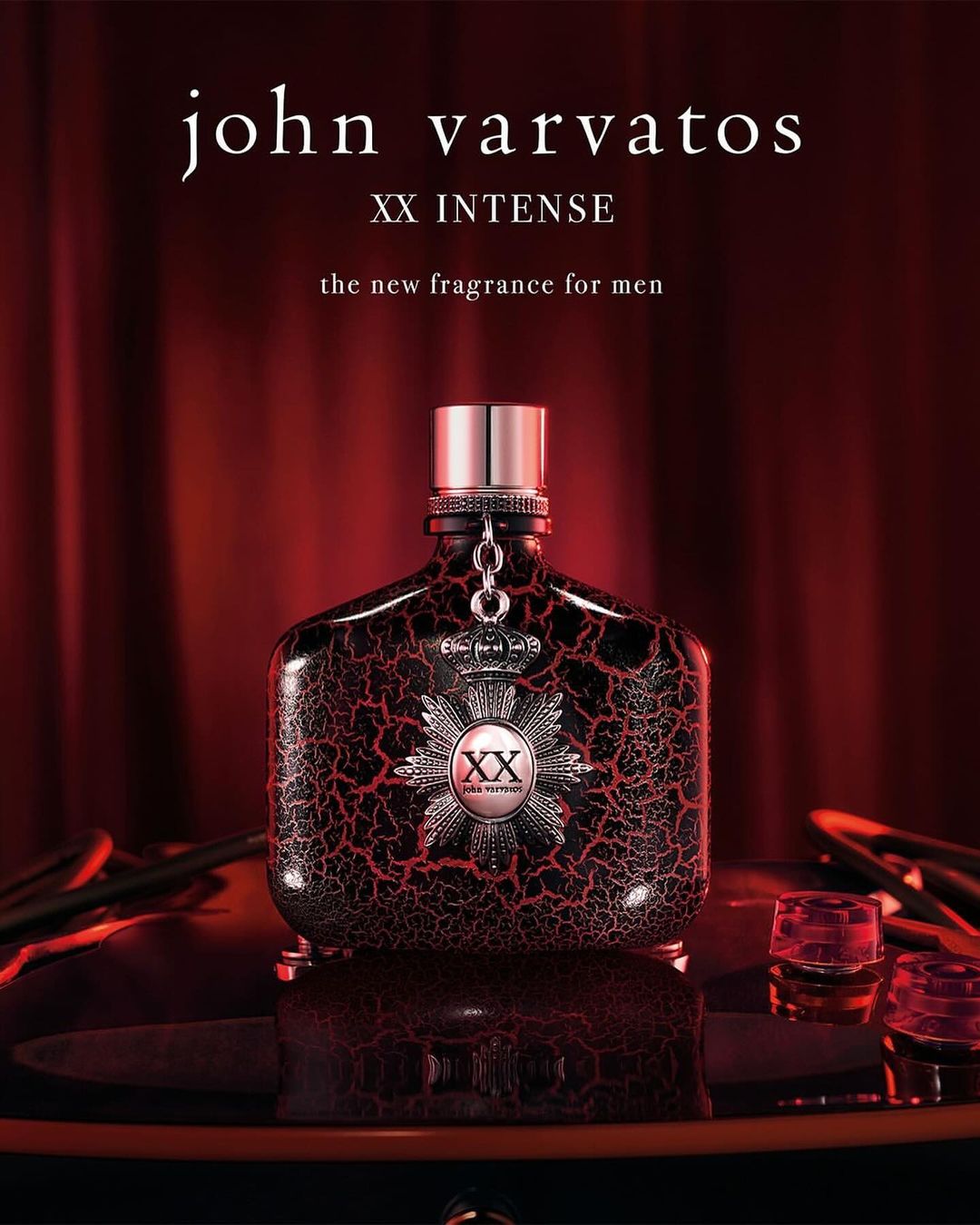 John Varvatos XX Intense fragrance bottle on a reflective surface, with two glasses and a red curtain backdrop. Product shot.
