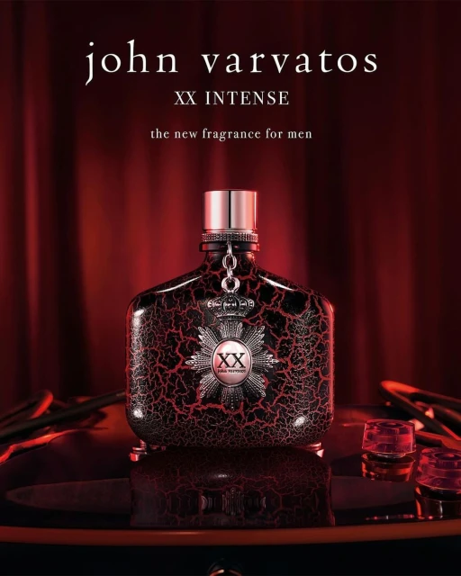 John Varvatos XX Intense fragrance bottle on a reflective surface, with two glasses and a red curtain backdrop. Product shot.