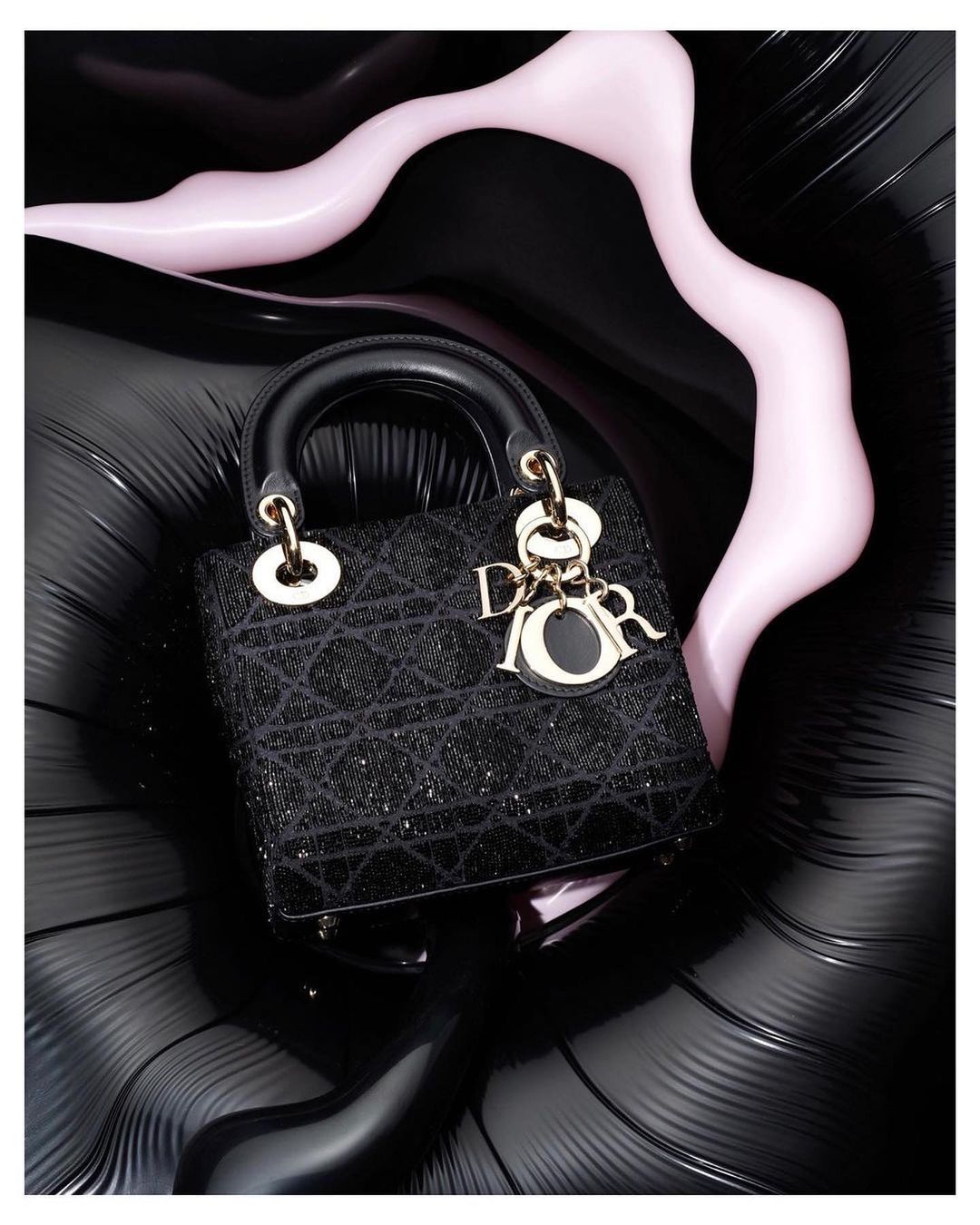 Fashion shot of a black Lady Dior handbag with gold hardware on a black surface with a pink abstract shape in the background.