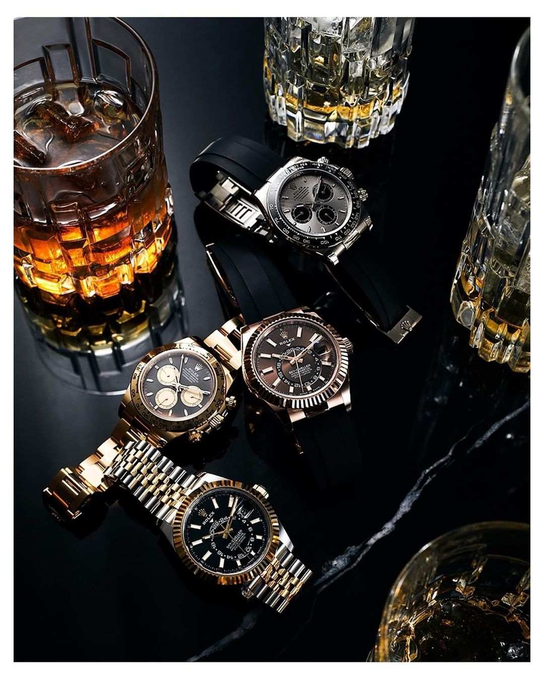 Four luxury watches with gold, silver, and black bands are displayed on a black surface with cut crystal whiskey glasses, indoor lighting.