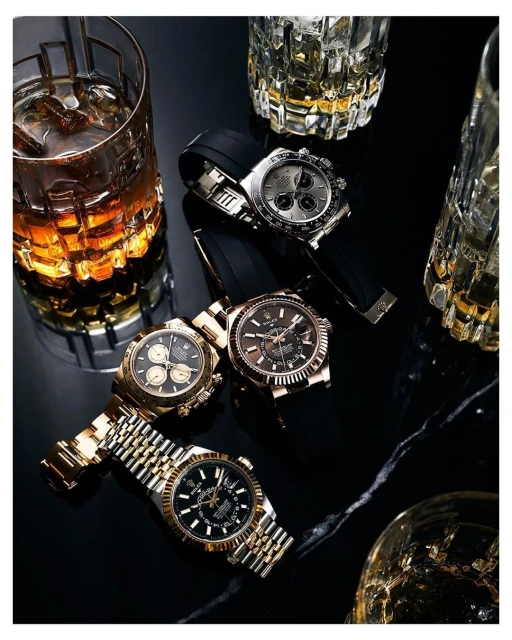 Four luxury watches with gold, silver, and black bands are displayed on a black surface with cut crystal whiskey glasses, indoor lighting.
