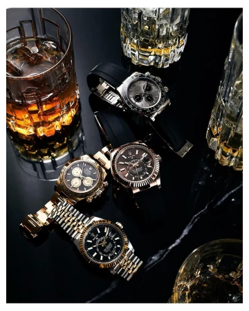 Four luxury watches with gold, silver, and black bands are displayed on a black surface with cut crystal whiskey glasses, indoor lighting.