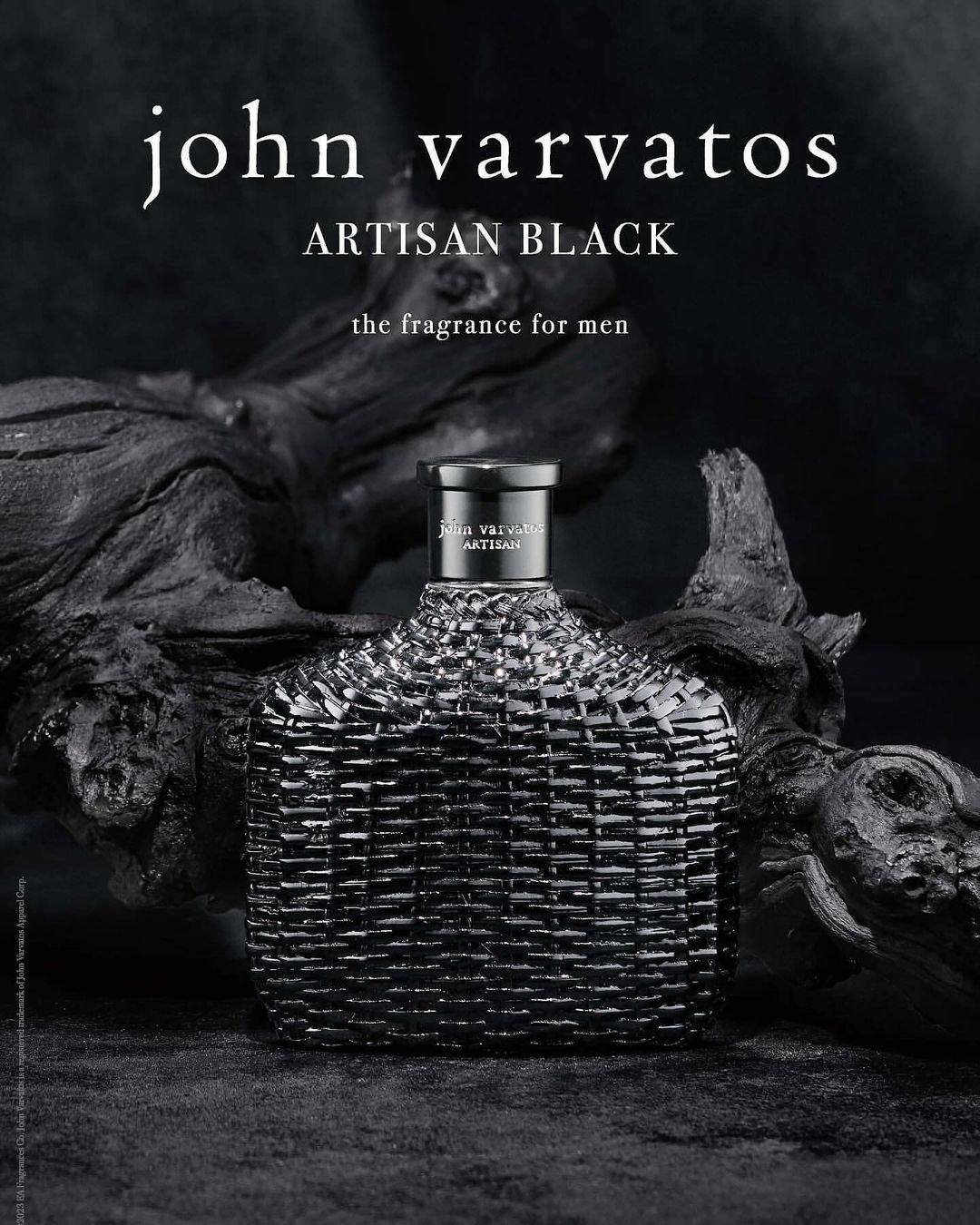 John Varvatos Artisan Black fragrance bottle close-up, displayed on a dark wooden surface against a blurred black background.