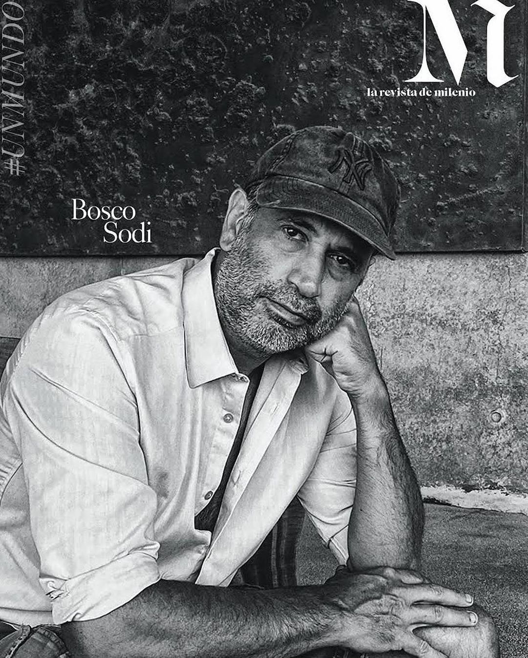 Black and white portrait of Bosco Sodi wearing a baseball cap and button-down shirt, posing in front of a textured background.