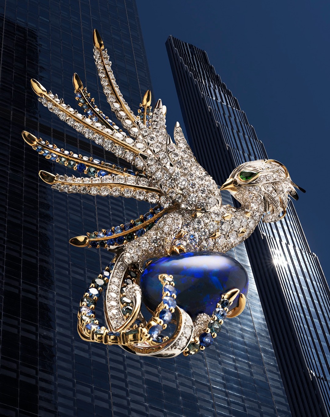 Detailed jewelry brooch in the form of an eagle with wings spread, perched on a dark blue opal, set against a blurred building background.