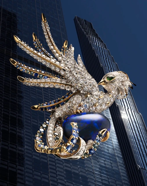 Detailed jewelry brooch in the form of an eagle with wings spread, perched on a dark blue opal, set against a blurred building background.