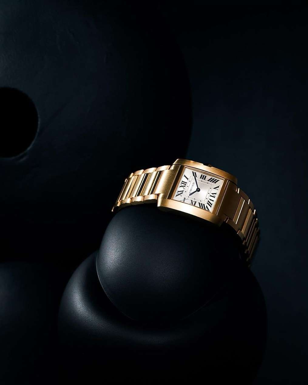 Elegant gold wristwatch with a white face displayed against black abstract forms creating a minimalist and luxurious product shot.