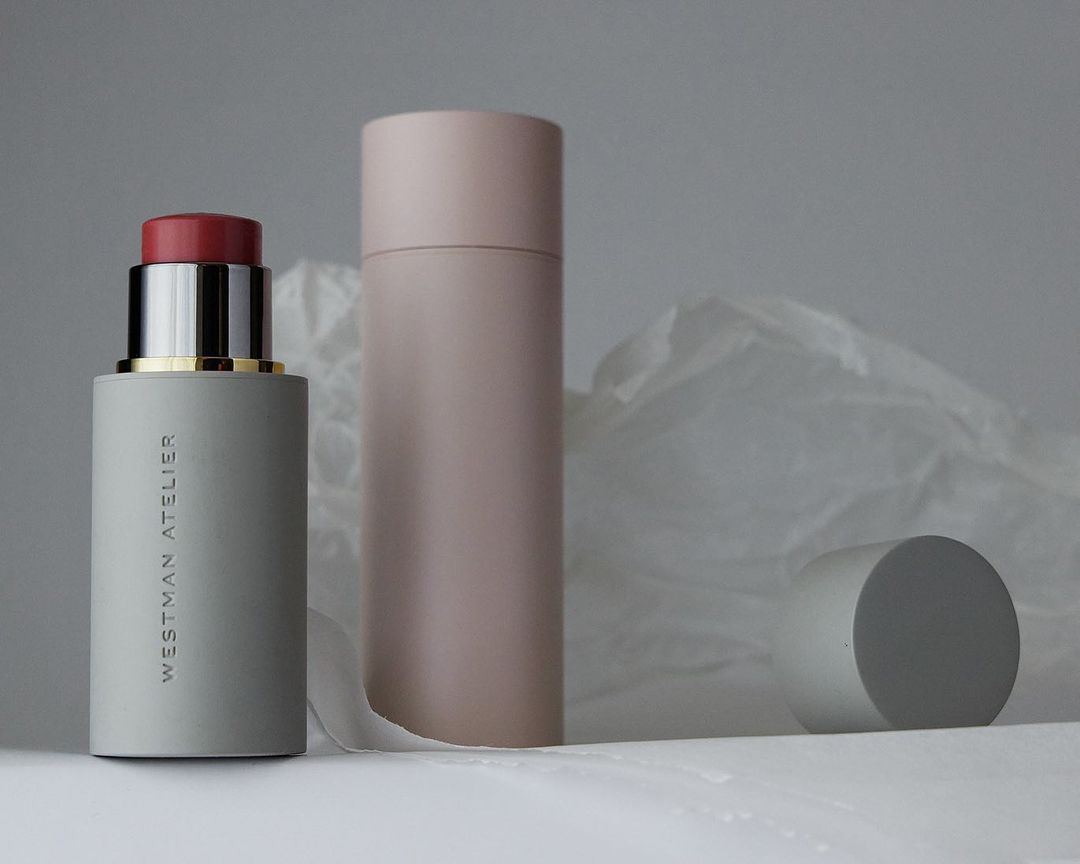 Cosmetic still life: blush stick with cap off, blush tube, and cylinder against a white and gray backdrop and folded paper.
