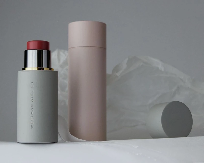 Cosmetic still life: blush stick with cap off, blush tube, and cylinder against a white and gray backdrop and folded paper.