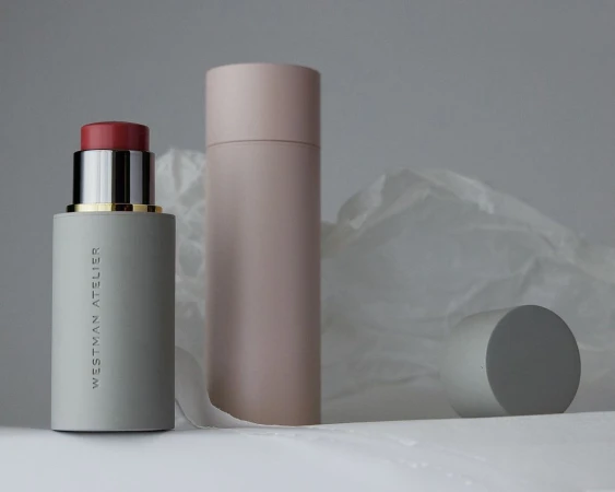 Cosmetic still life: blush stick with cap off, blush tube, and cylinder against a white and gray backdrop and folded paper.