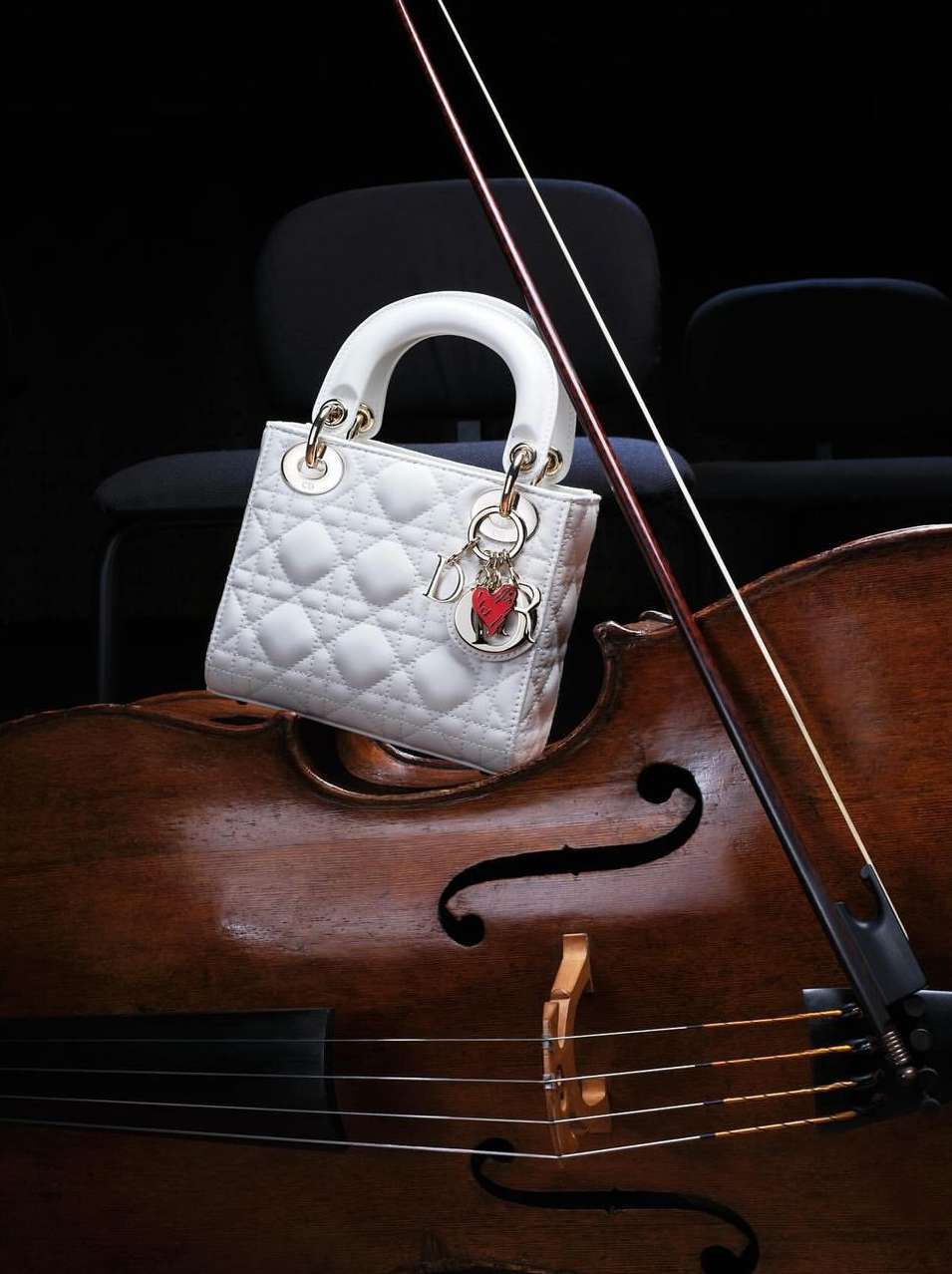 White quilted Lady Dior handbag with gold hardware and charms displayed on the body of a cello in a dark, indoor setting. 