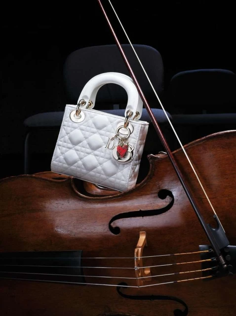 White quilted Lady Dior handbag with gold hardware and charms displayed on the body of a cello in a dark, indoor setting. 