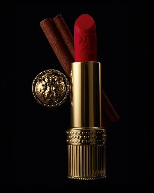 Close-up studio shot of red lipstick with gold tube, decorative gold medallion, and cinnamon sticks against a black background.