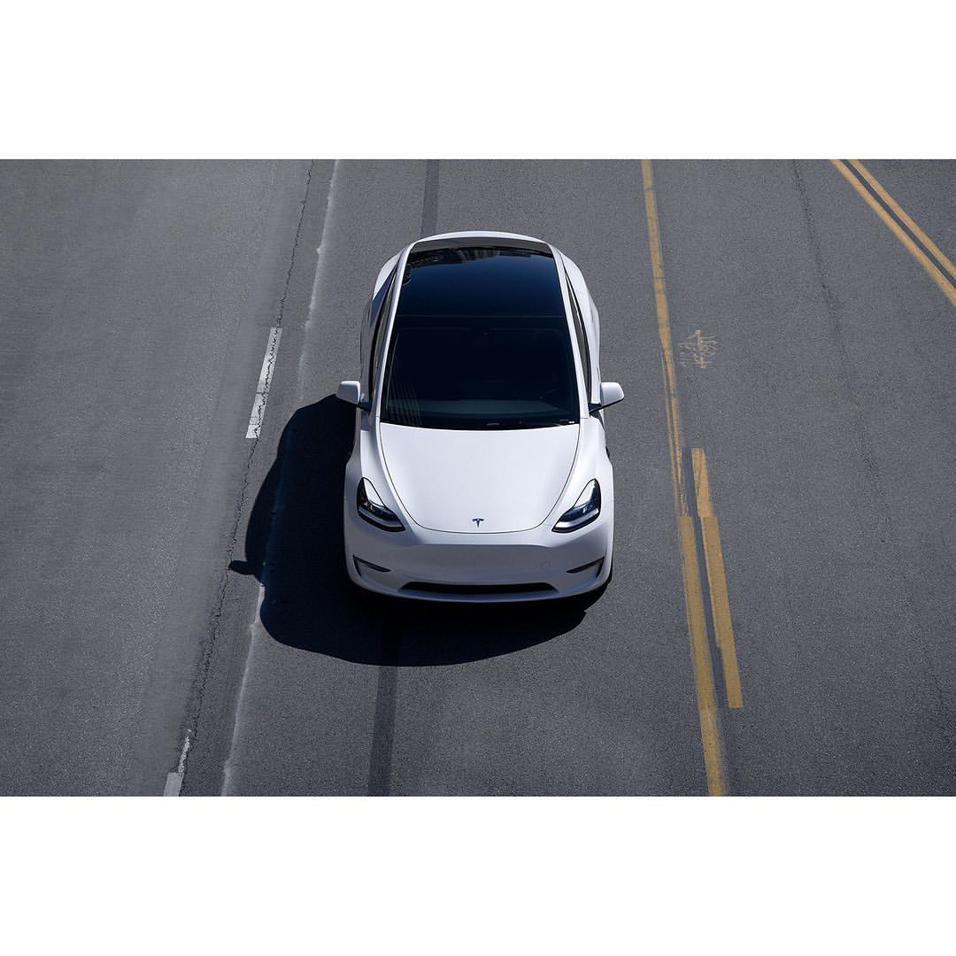 Aerial view of a white Tesla Model Y electric SUV on grey asphalt roadway with yellow painted lane divider, captured in daylight.