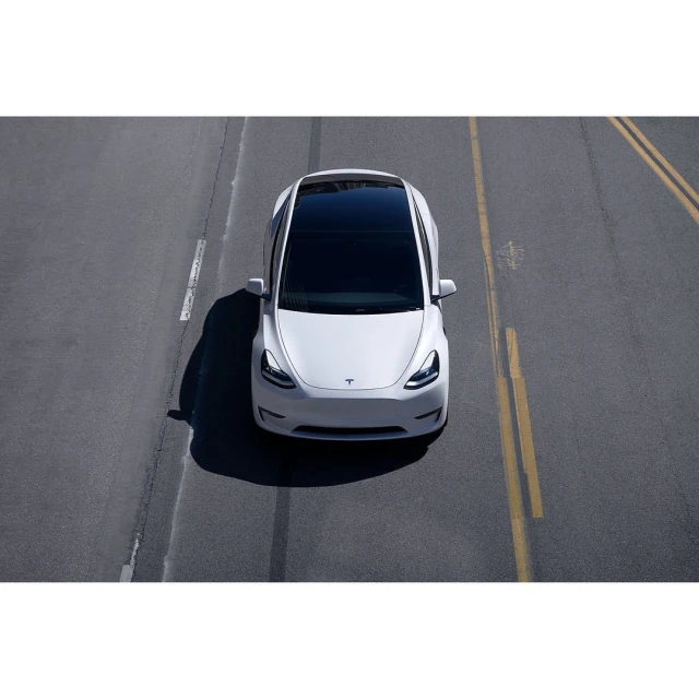 Aerial view of a white Tesla Model Y electric SUV on grey asphalt roadway with yellow painted lane divider, captured in daylight.