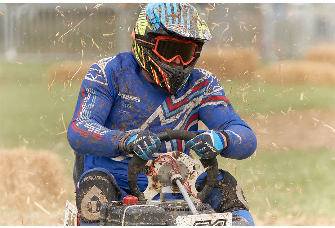 Action shot of a racer in full protective gear and helmet racing on a lawnmower on a dirt track outdoors, with hay bales in the background.