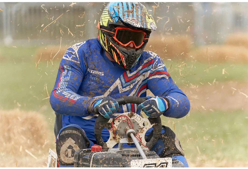 Action shot of a racer in full protective gear and helmet racing on a lawnmower on a dirt track outdoors, with hay bales in the background.