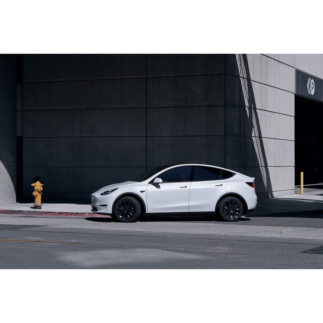 White Tesla Model Y parked on a city street with modern building and fire hydrant in the background, offering a sleek and urban aesthetic.