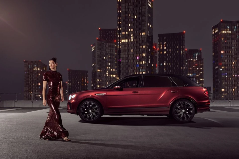 Woman in sequined gown poses next to a red SUV on a rooftop at night with city buildings visible in the background.