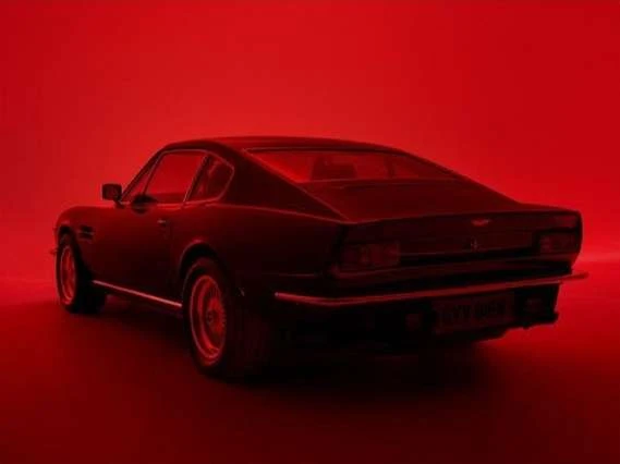 Rear quarter view of a classic black Aston Martin DBS on display, strongly lit with red studio lights, showcasing its iconic design.