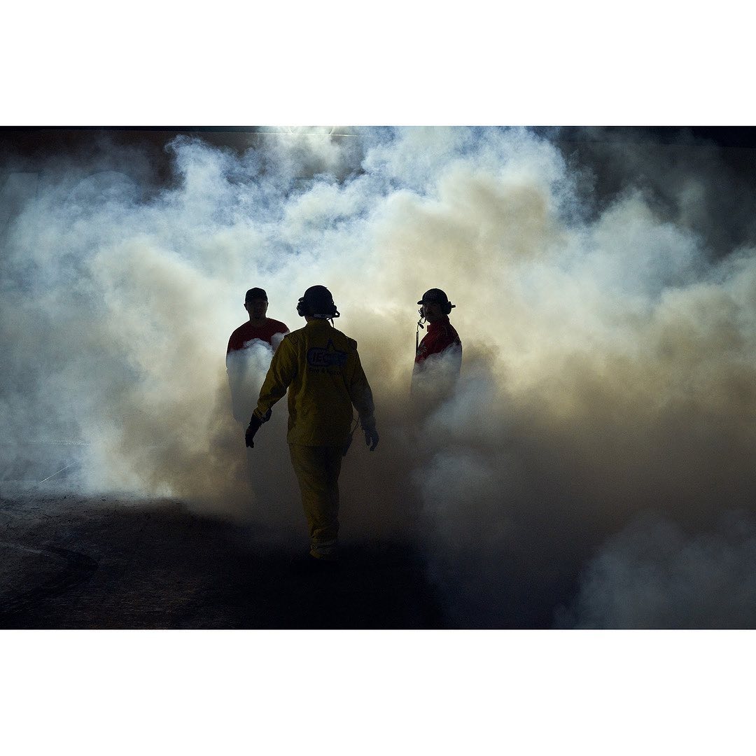 Three people in protective suits walk through thick smoke or fog in a dimly lit environment, creating a dramatic, obscured scene.