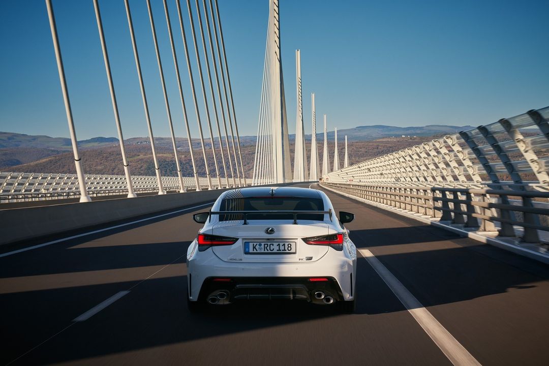 A white Lexus RC is driving on a modern bridge with support pillars, showcasing automotive design and the scenic landscape in the background.