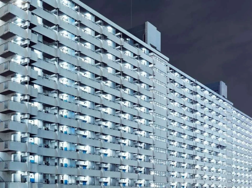 Exterior shot of a large grey apartment building at night, showcasing numerous illuminated windows and balconies against a dark sky.