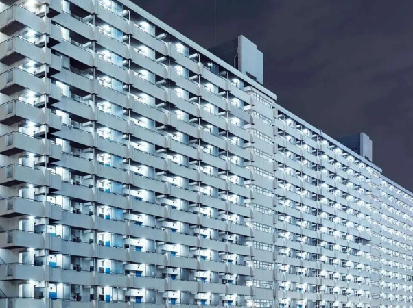 Exterior shot of a large grey apartment building at night, showcasing numerous illuminated windows and balconies against a dark sky.