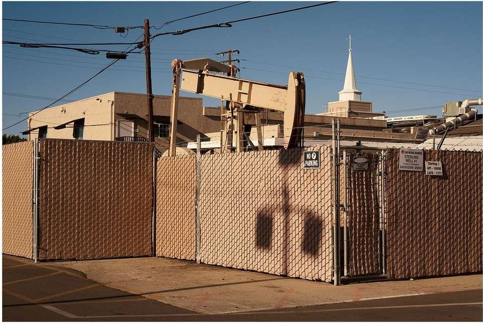Oil pump jack behind a chain-link fence in an urban setting, with a church steeple visible in the background against a clear blue sky.