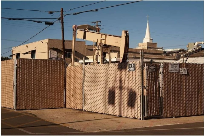Oil pump jack behind a chain-link fence in an urban setting, with a church steeple visible in the background against a clear blue sky.