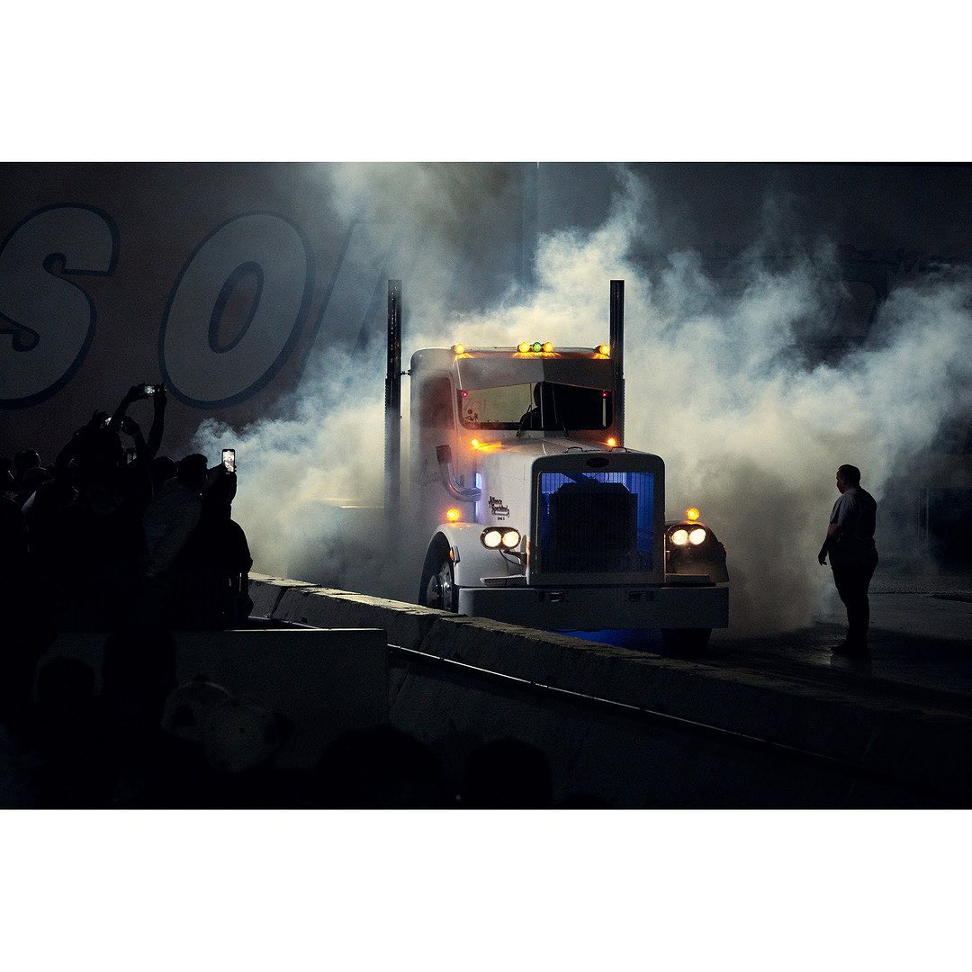 White semi-truck enveloped in smoke, stage lights illuminate scene, crowd silhouetted on left, adult male standing on right.