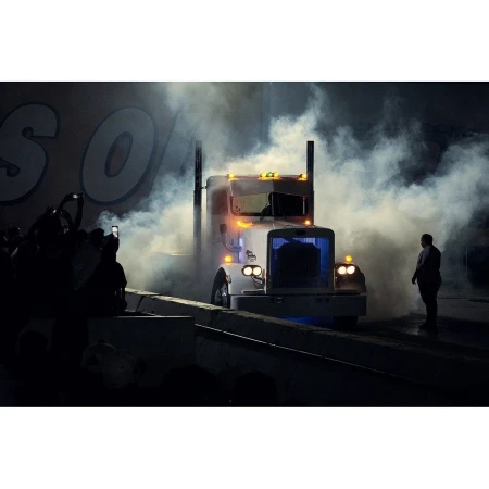 White semi-truck enveloped in smoke, stage lights illuminate scene, crowd silhouetted on left, adult male standing on right.