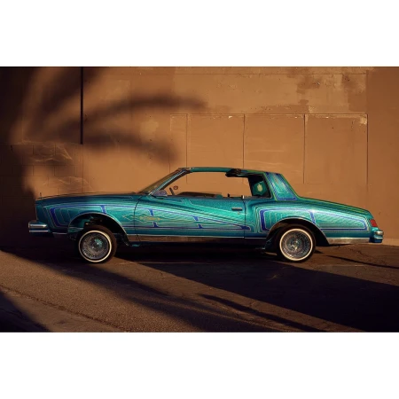 Customized teal lowrider car parked on a street with a shadow from a tree cast on the wall behind it in the daytime.